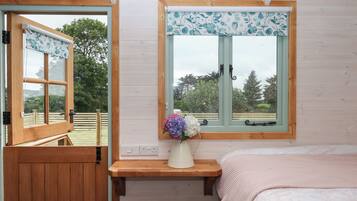 A bedroom with a wooden bed, a nightstand, a window with a floral curtain, and a view of a wooden deck and trees.