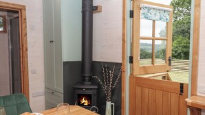 A cozy room with a wood-burning stove, a dining table with breakfast items, and a view through a window. - Lottie's Hut (Okehampton)