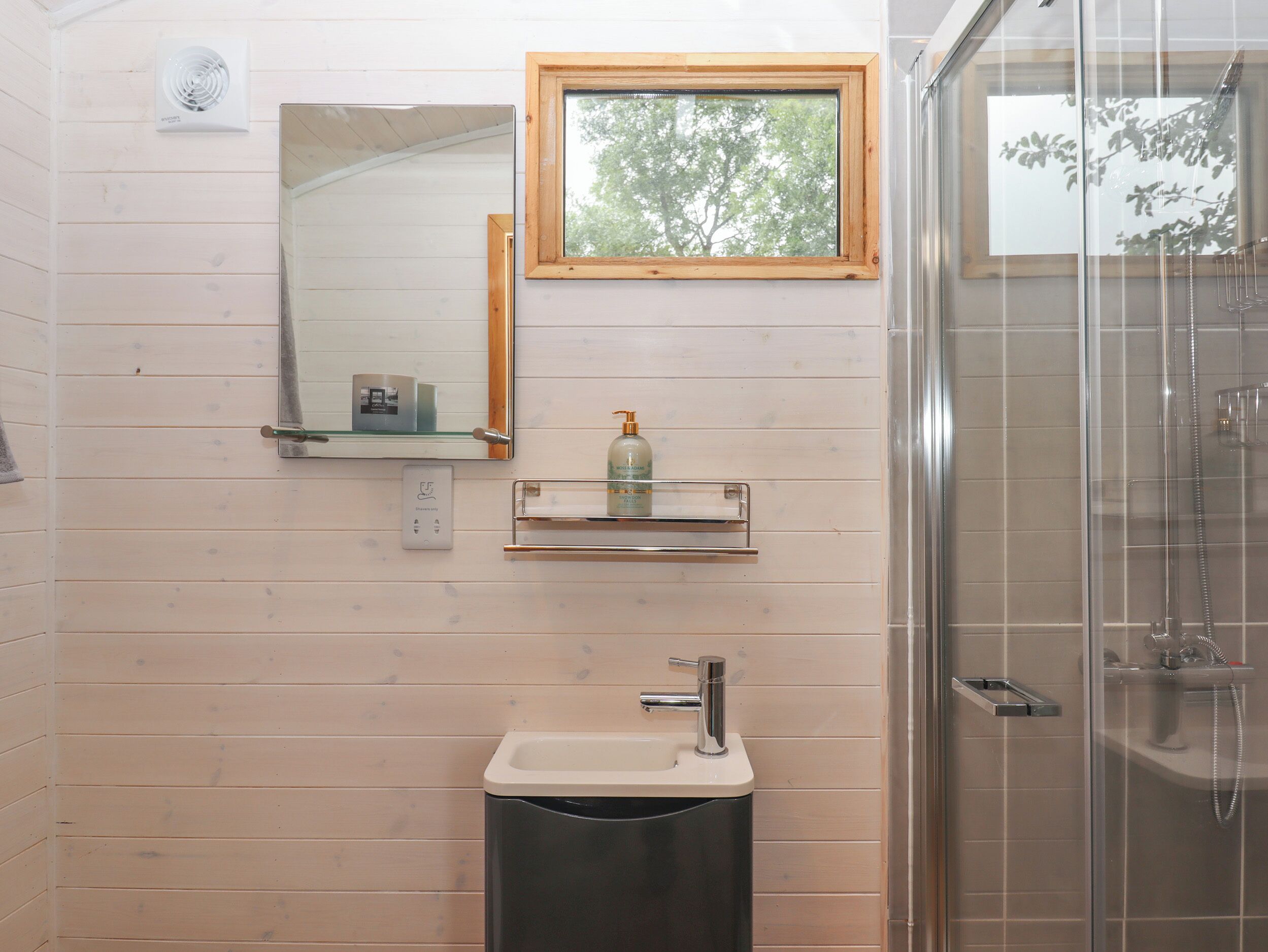 Une salle de bain avec un mur lambrissé en bois, un miroir, un lavabo, une douche en verre et une fenêtre donnant sur des arbres.