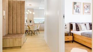 A modern interior with a wooden bench, a dining area with chairs, a kitchen in the background, and a bed with pillows and a nightstand. - Lumina glamour 3 bedroom apartment (Lodz)