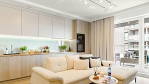 A modern living room with a beige sofa, a small coffee table, and a kitchen area with wooden cabinets and a built-in oven. - Lumina glamour 3 bedroom apartment (Lodz)