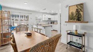 Una cocina moderna con armarios blancos, una isla central y un comedor con mesa de madera y sillas de mimbre.