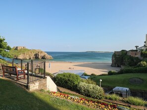 A coastal view with a beach, a castle-like structure, and a garden with colorful flowers. - Flat 1 (Tenby)