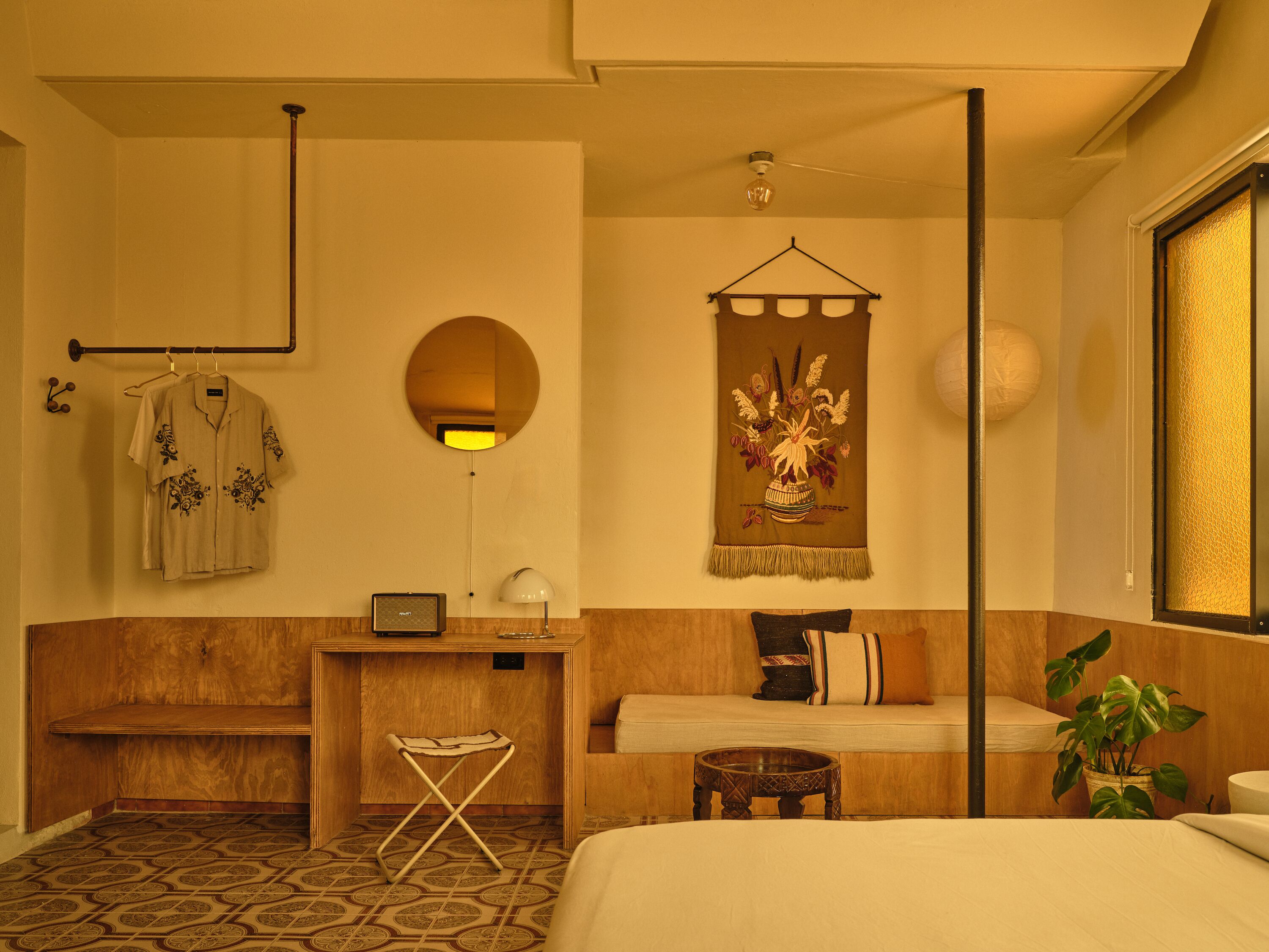 A hotel room with a bed, a wooden bench, a small table, a hanging mirror, a wall-mounted lamp, a wall decoration, and a potted plant.