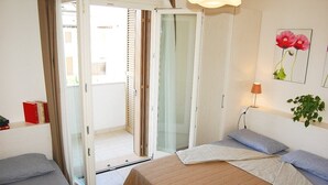 Une chambre avec deux lits, une porte-fenêtre coulissante donnant sur un balcon, une lampe de chevet et un mur orné d’un tableau représentant des fleurs.