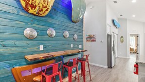 A bar area with a wooden counter and red bar stools, a blue wooden wall with decorative plates, and a kitchen in the background.