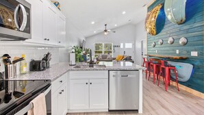 A modern kitchen with white cabinets, a stainless steel dishwasher, and a granite countertop. There is a dining area with red chairs and a blue wall with decorative plates.