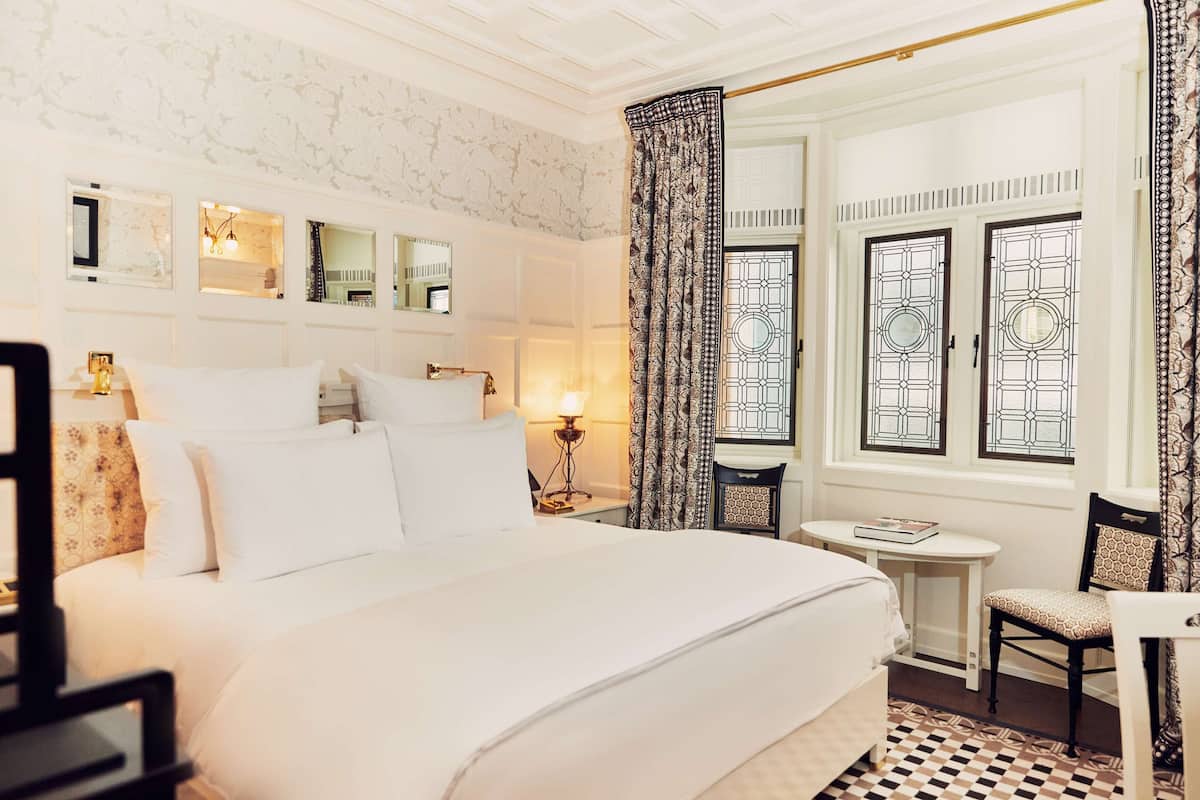 A hotel room with a large bed, a small table with a chair, a mirror, and a window with decorative glass panes.