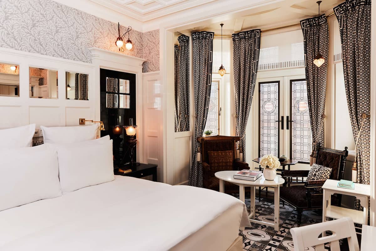 A hotel room with a large bed, a small table with a vase, a chair, and a desk. There are curtains on the windows and a patterned floor.