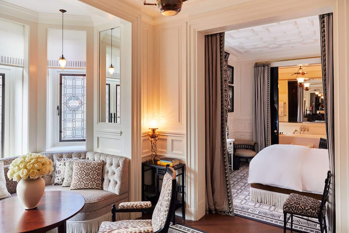 A luxurious hotel room with a large bed, a sitting area with a sofa and chairs, a small table with a vase of flowers, and a mirror.