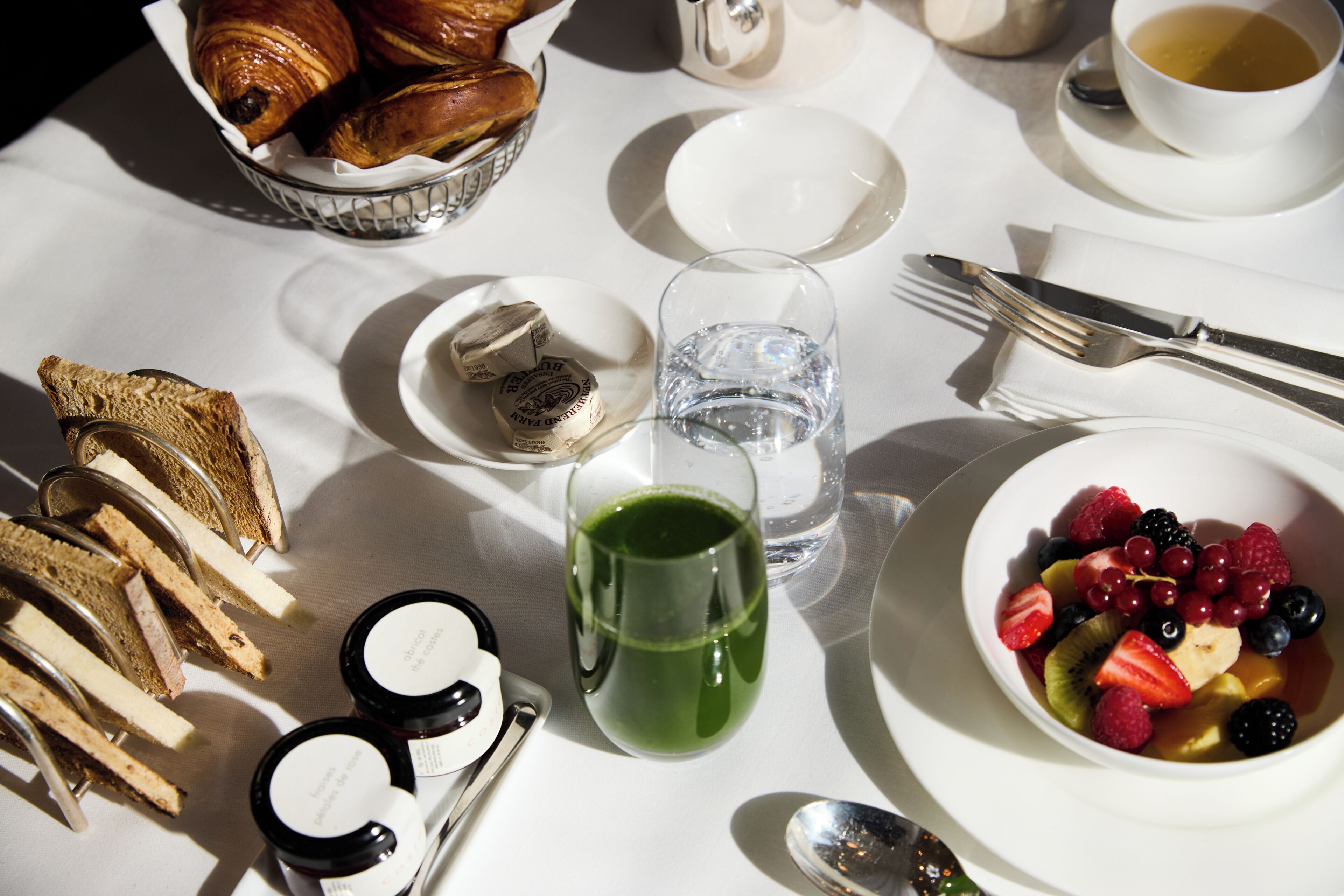 Een ontbijtje met croissants, fruit, jam en een groen drankje op een tafel.