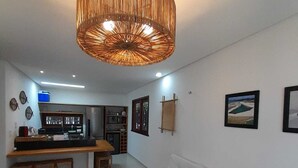 A modern living room with a white sofa, a wooden bench, a woven ceiling light fixture, and a kitchen area with a refrigerator and a sink.
