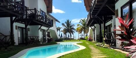 A pool area with a wooden deck, a grassy lawn, and a thatched-roof building.