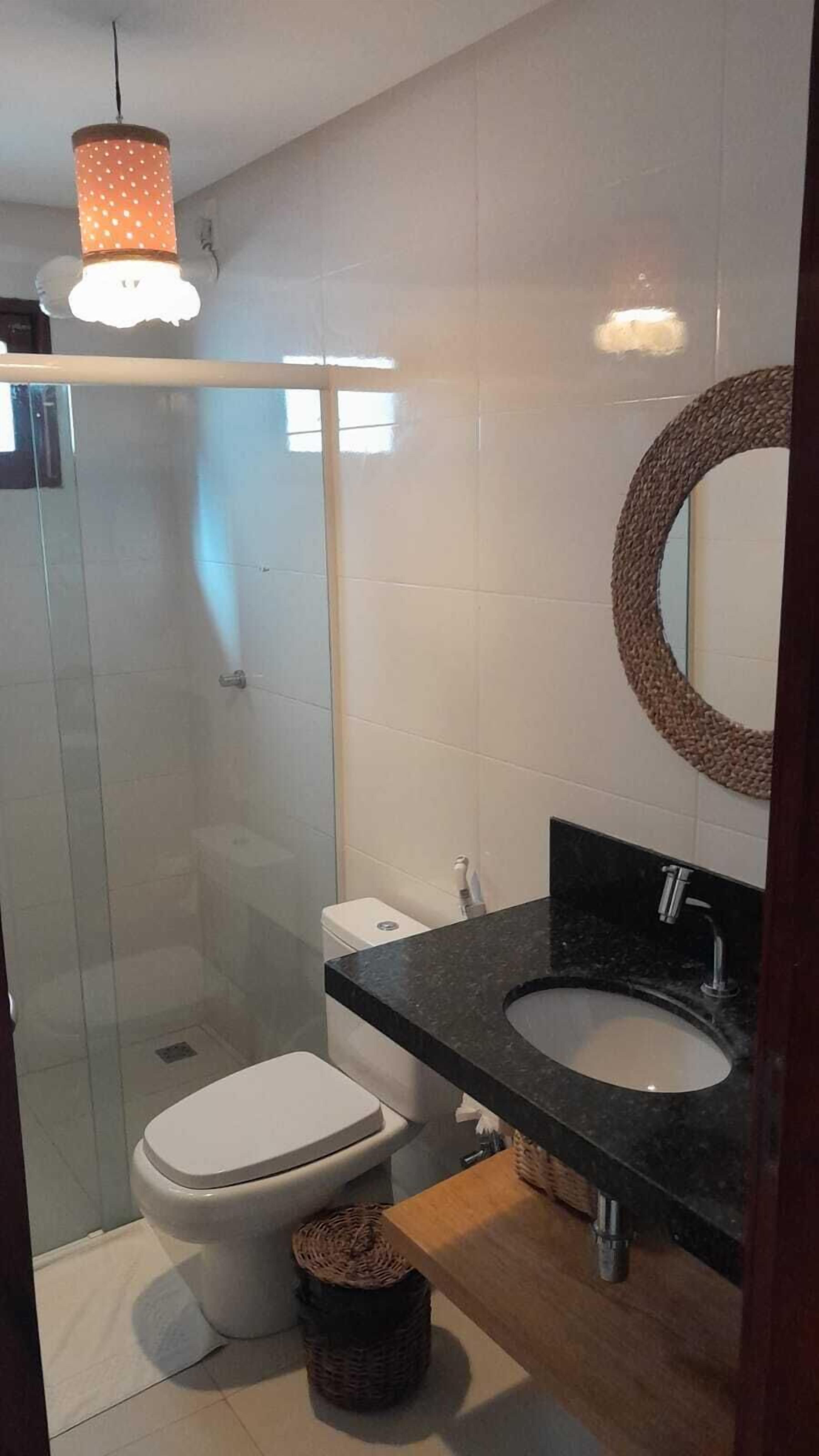 A bathroom with a glass shower enclosure, a toilet, a sink with a dark countertop, and a round mirror with a woven frame.