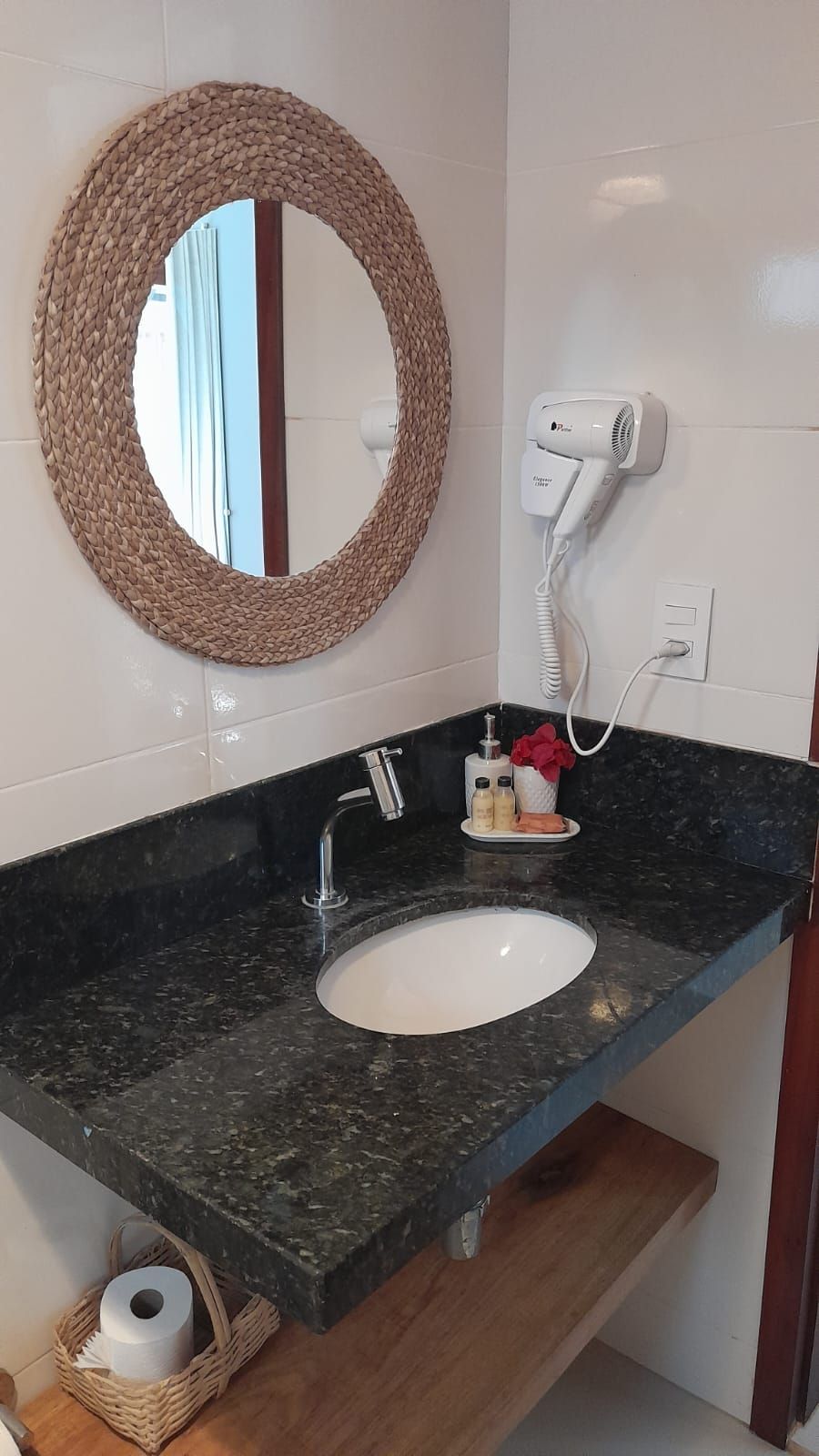A bathroom with a round woven mirror, a white sink, a hairdryer, and a small basket with a roll of toilet paper.