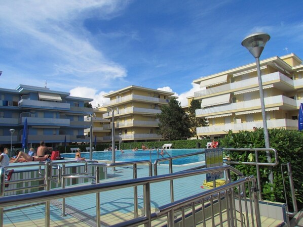 A swimming pool area with multiple balconies, a poolside lounge, and a clear sky. - Homely Apartment With Shared Pools - Beahost (San Michele al Tagliamento)