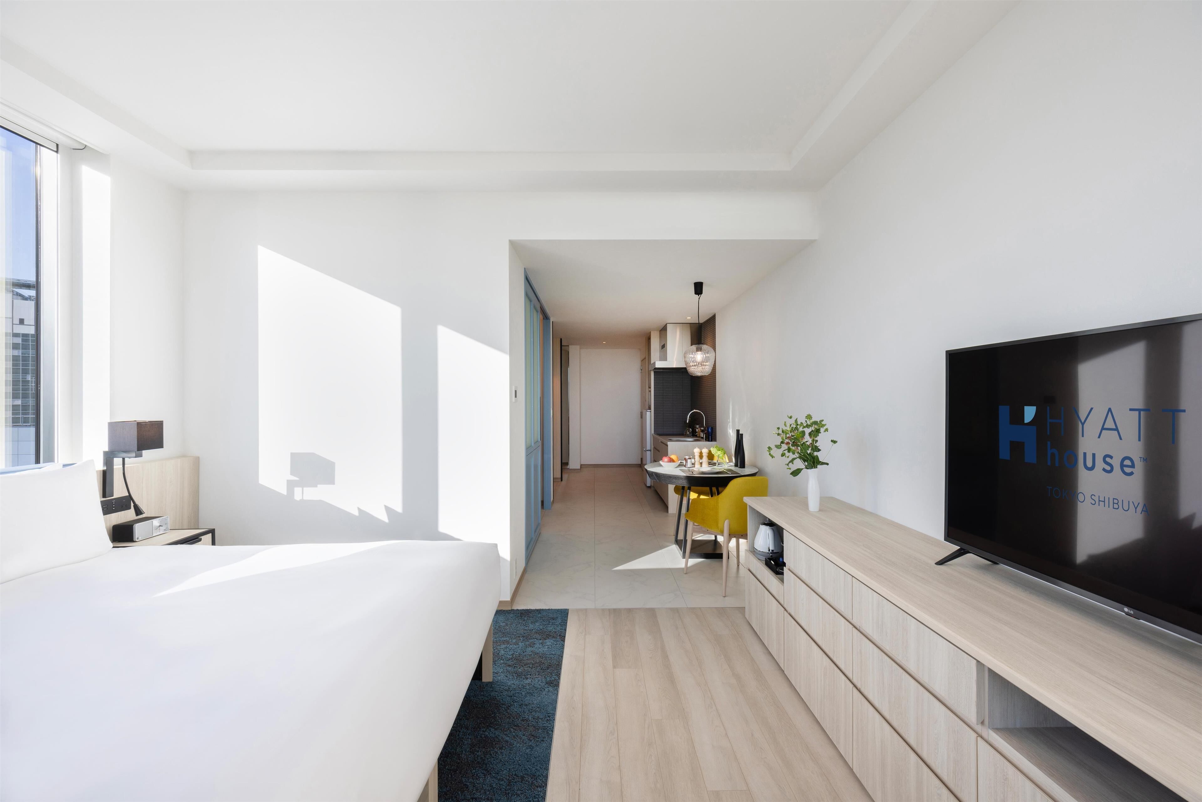 A modern hotel room with a large bed, a flat-screen TV, a dining area with a yellow chair, and a kitchenette.
