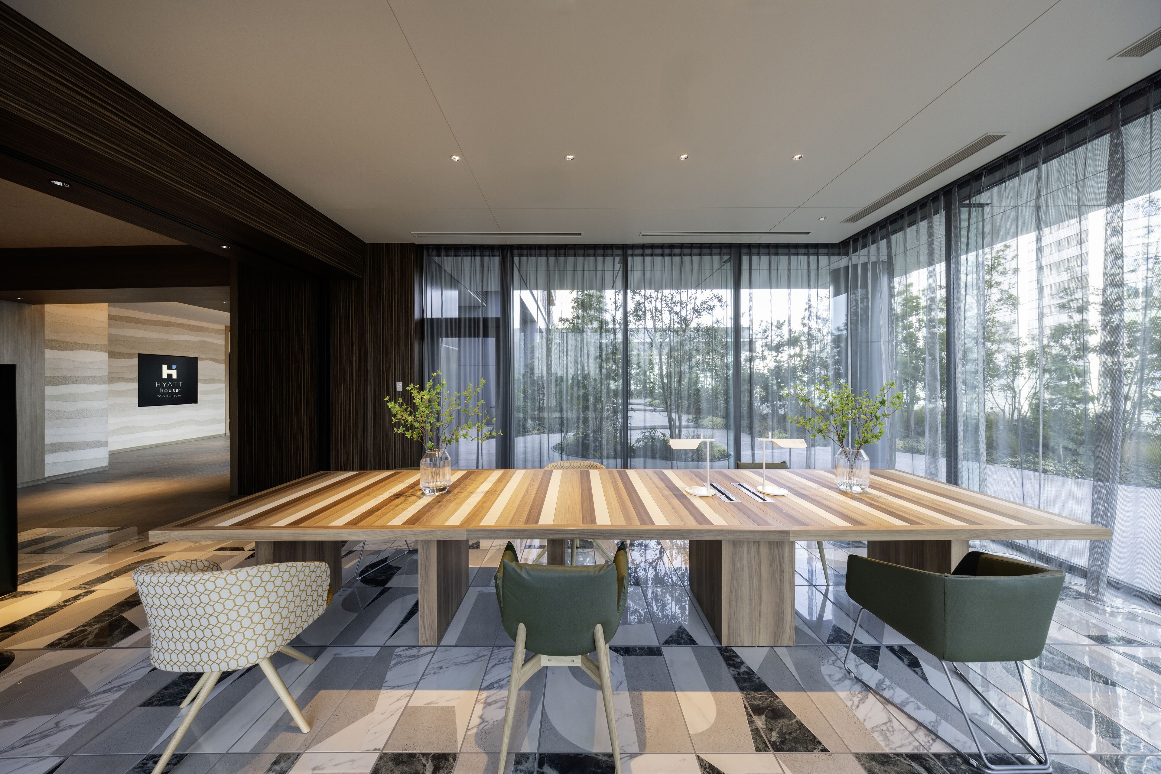 A modern conference room with a large wooden table, two green chairs, and a glass partition.
