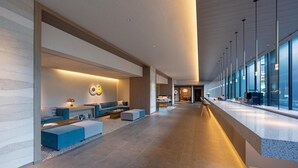 Un hall d’hôtel moderne doté d’un long comptoir d’accueil, d’un espace salon avec des canapés et des poufs, et de grandes fenêtres offrant une vue sur la ville.