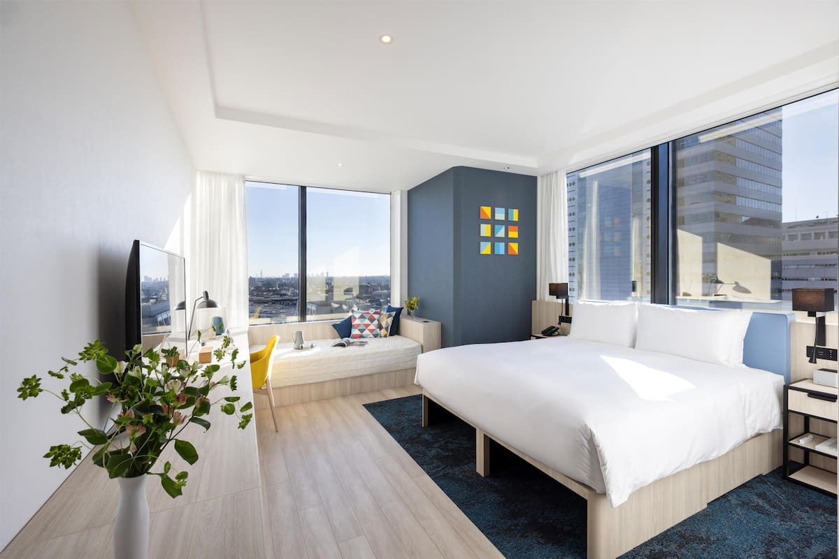 A modern hotel room with a large bed, a seating area with a view, and a cityscape visible through the windows.