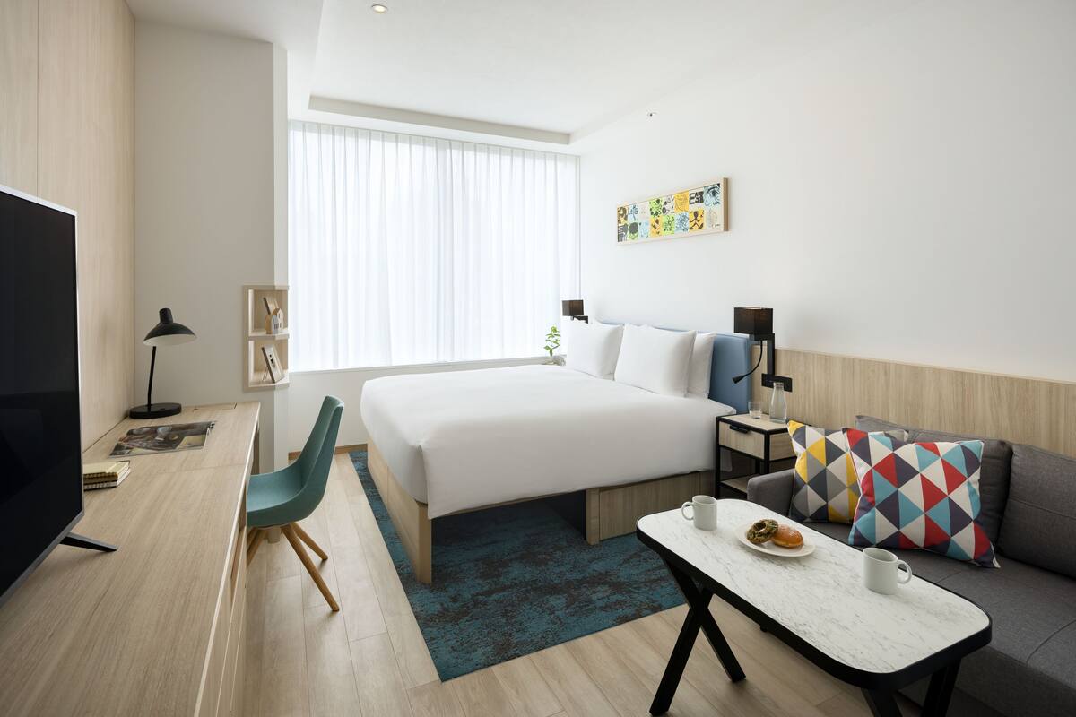 A modern hotel room with a large bed, a desk with a chair, a sofa, and a television.