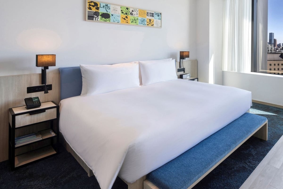 A hotel room with a large bed, two bedside tables, a nightstand, a bench, a wall art piece, and a window with a city view.