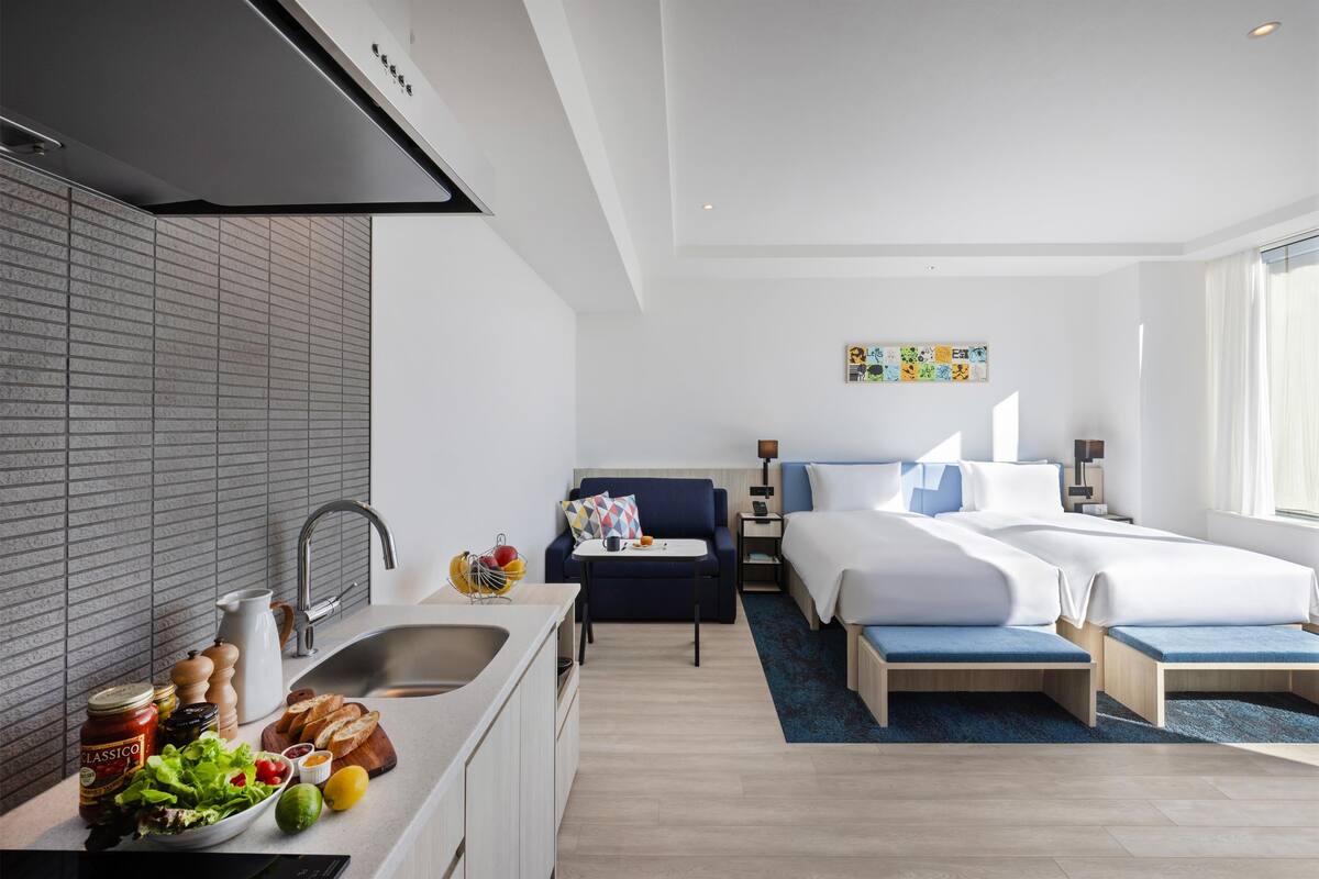 A modern hotel room with a kitchenette, a bed with blue bedding, a small seating area, and a view of the city.