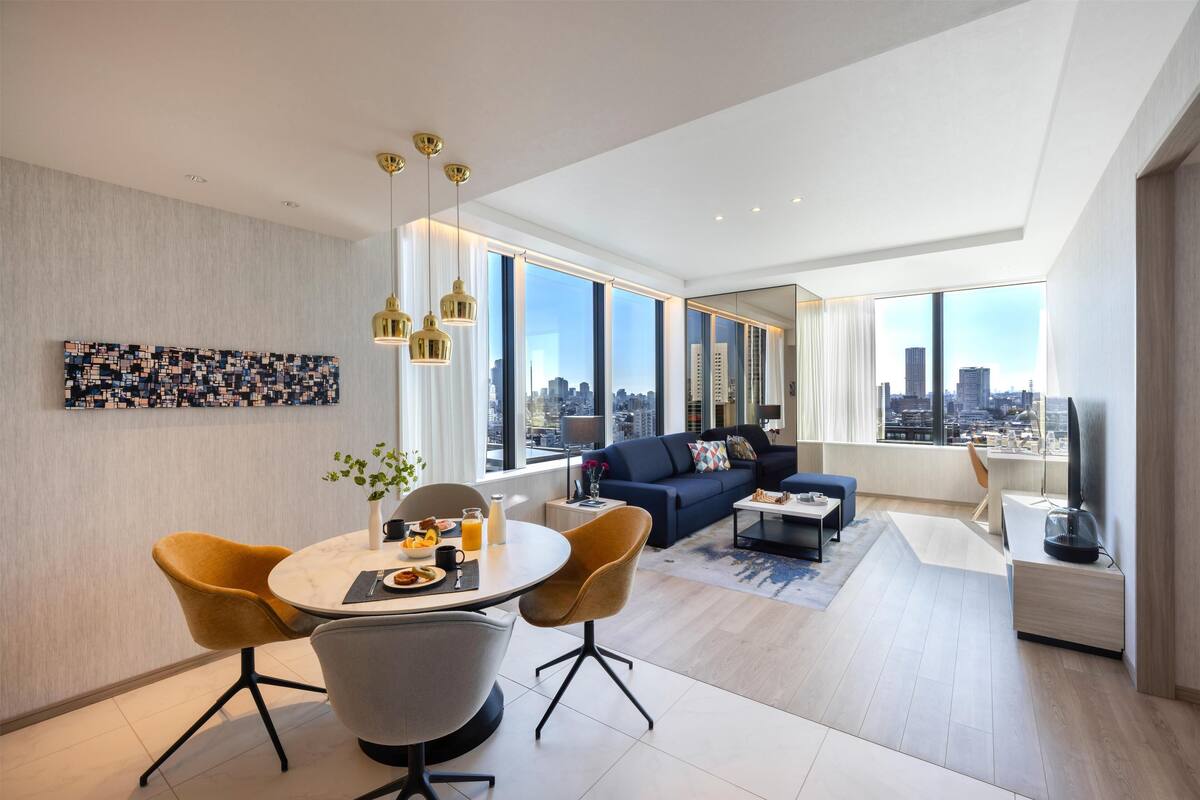A modern living room with a dining area, large windows with a city view, and a sofa with decorative pillows.