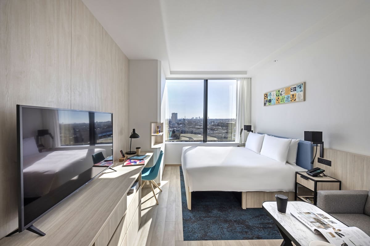 A modern hotel room with a large bed, a desk, a flat-screen TV, and a large window offering a city view.