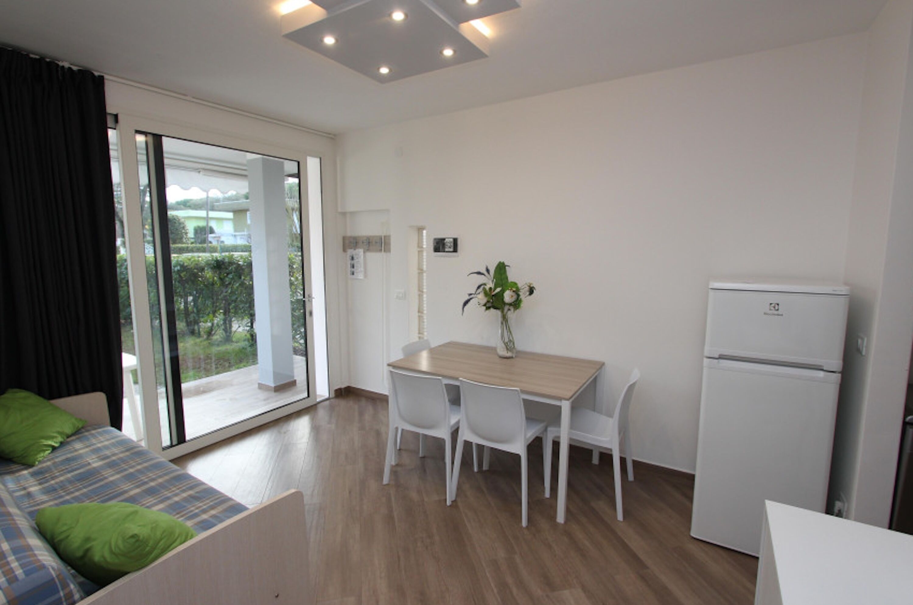 A modern room with a wooden dining table, white chairs, a small refrigerator, and a bed with green pillows.