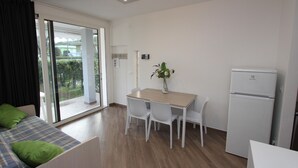 Une chambre moderne avec une table à manger en bois, des chaises blanches, un petit réfrigérateur et un lit agrémenté d’oreillers verts.