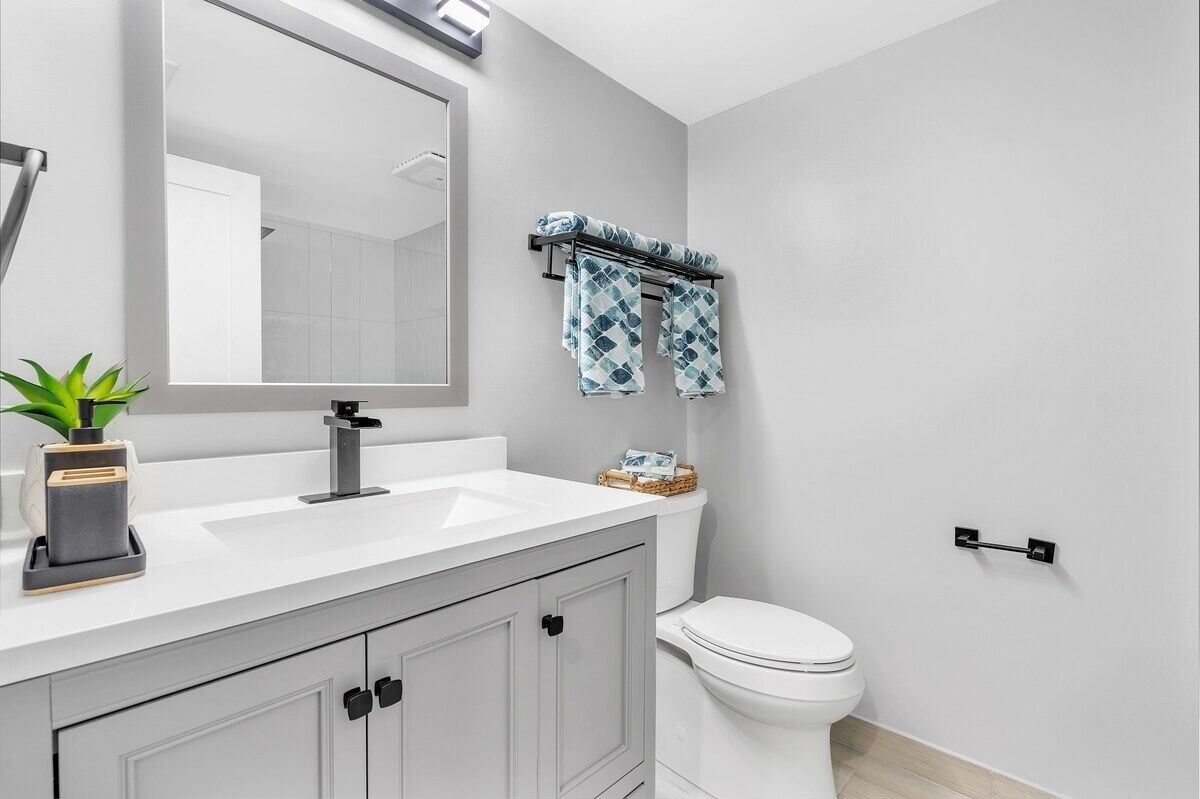 A modern bathroom with a white sink, a large mirror, a towel rack with patterned towels, a toilet, and a small plant on the counter.