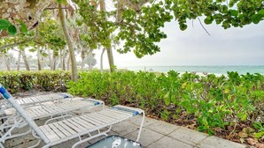 A beachside lounge area with white lounge chairs and a glass table, surrounded by greenery and overlooking the sea.