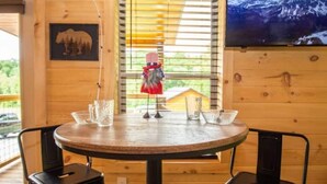 A wooden interior with a round table, two chairs, a bear picture, a small figurine, and a window with blinds. - Couple's Retreat: HotTub-FirePit-Grill-Pets-Wifi (Sevierville)