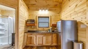 A compact kitchen with wooden cabinets, a stainless steel refrigerator, a microwave, and a window with blinds. - Couple's Retreat: HotTub-FirePit-Grill-Pets-Wifi (Sevierville)
