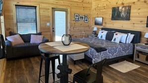 A cozy log cabin room with a bed, a sofa, a dining table, and a TV mounted on the wall. - Couple's Retreat: HotTub-FirePit-Grill-Pets-Wifi (Sevierville)