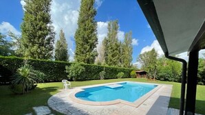A rectangular swimming pool with a tiled deck, surrounded by a well-maintained lawn and tall trees. - Charming Villa With Pool in Albarella (Rosolina)