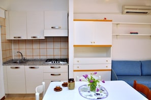 A compact kitchen with white cabinets, a gas stove, and a sink. There's a dining table with a vase of flowers and a plate. A blue sofa is against the wall.