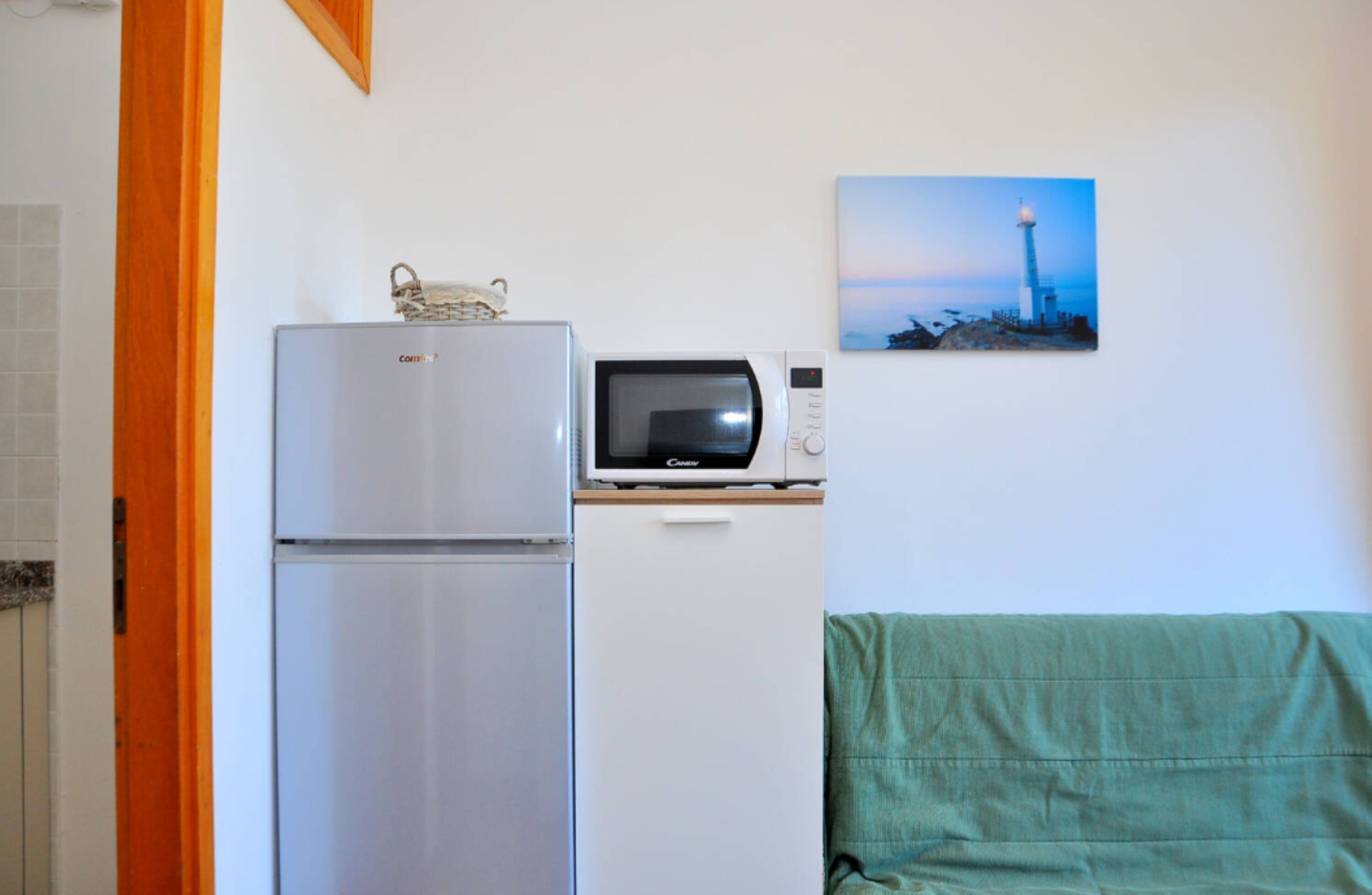 A compact kitchenette with a refrigerator, microwave, and a small cabinet. A painting of a lighthouse is on the wall.
