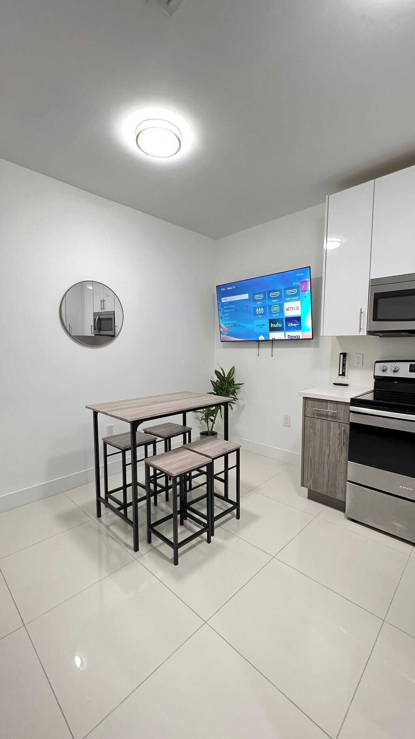 Una cocina moderna con armarios blancos, horno empotrado y microondas. Hay un espejo redondo en la pared y un televisor de pantalla plana colocado sobre una mesa de comedor de madera con bancos.