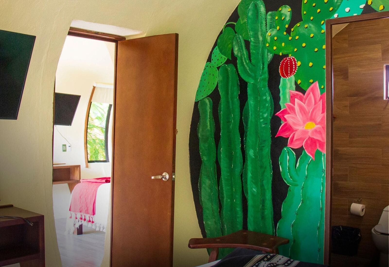 A room with a wooden door, a bed with a pink blanket, and a cactus painting on the wall.