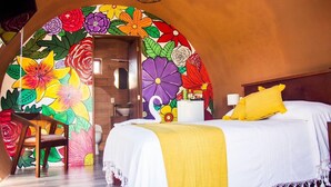 Un dormitorio con un mural floral colorido, una cama con ropa de cama blanca y amarilla, y un cabecero de madera.
