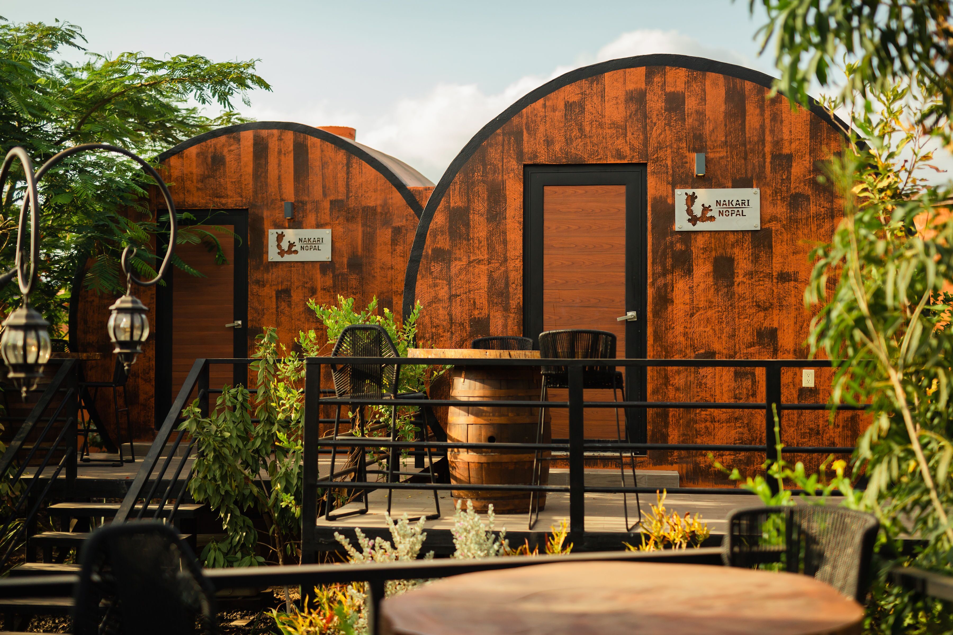 A rustic outdoor seating area with wooden tables and chairs, surrounded by metal railings and potted plants.