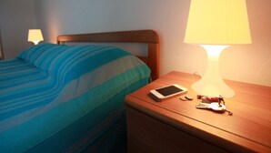 Een netjes opgemaakt bed met een blauw-groene gestreepte deken, een houten nachtkastje met een smartphone en sleutels, en een tafellamp die licht geeft.