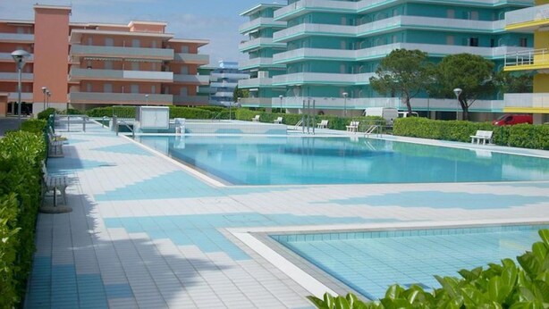 A swimming pool with a tiled deck, surrounded by greenery and modern buildings. - Sporty Residence With all you Need (San Michele al Tagliamento)