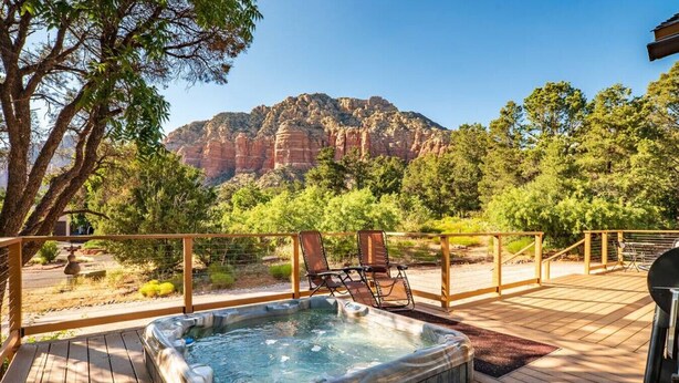 Un bain à remous sur une terrasse en bois, agrémentée de chaises, entouré d’arbres et offrant une vue sur la montagne.