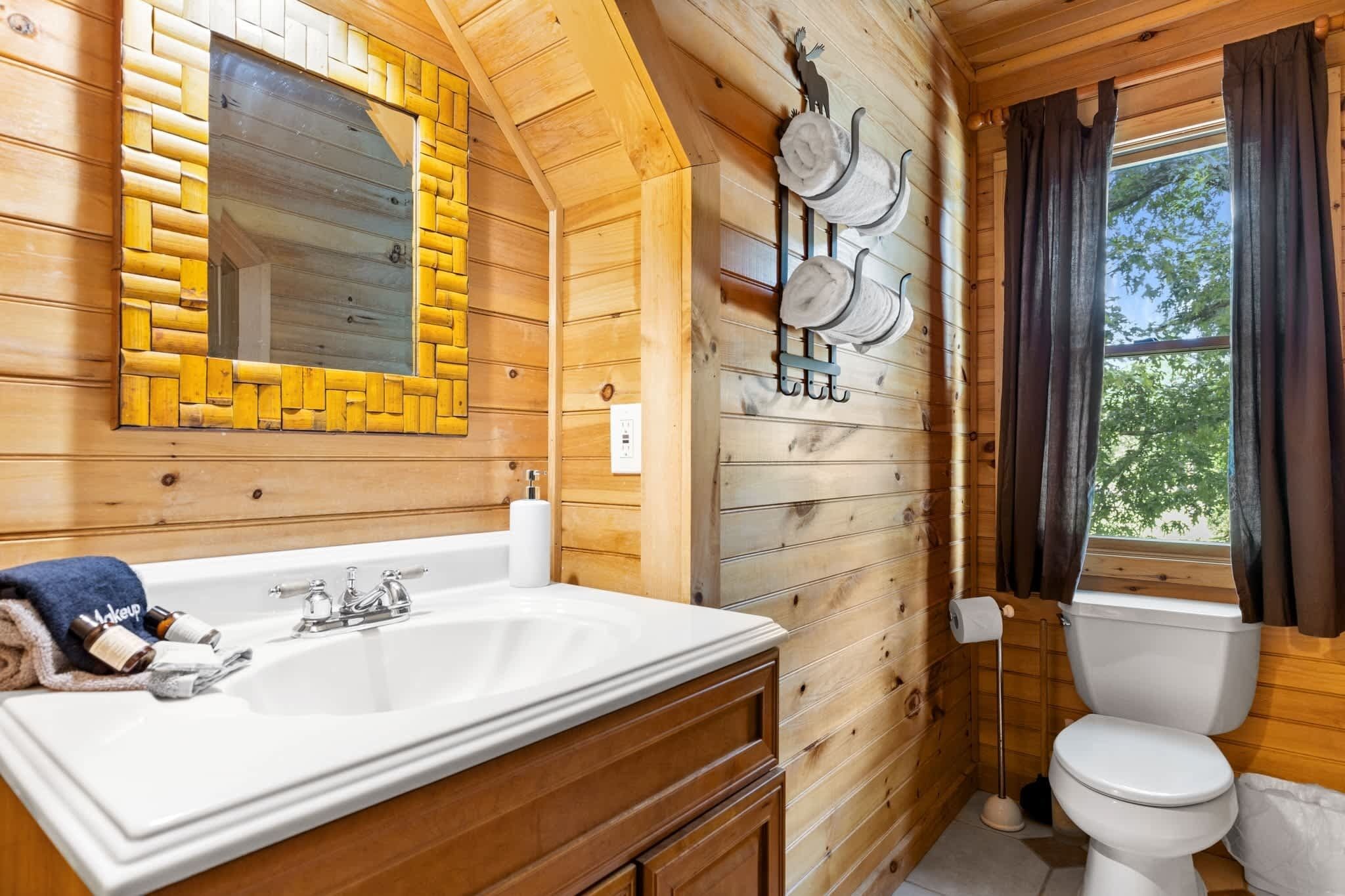 A wooden cabin bathroom with a sink, mirror, toilet, and window with curtains.