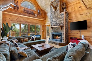 A cozy log cabin interior with a stone fireplace, a large sofa, and a television. - Lake Views | Hot Tub | Fire Pit | Micro Events (Morganton)