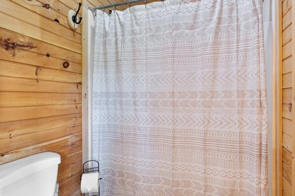 A bathroom with wooden walls, a white toilet, and a patterned shower curtain. - Lake Views | Hot Tub | Fire Pit | Micro Events (Morganton)