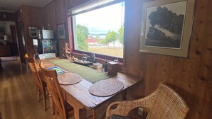 Ein Esszimmer mit Holzboden, einem großen Fenster, Korbstühlen und einem Tisch, der mit Untersetzern und einer dekorativen Buddha-Statue gedeckt ist.